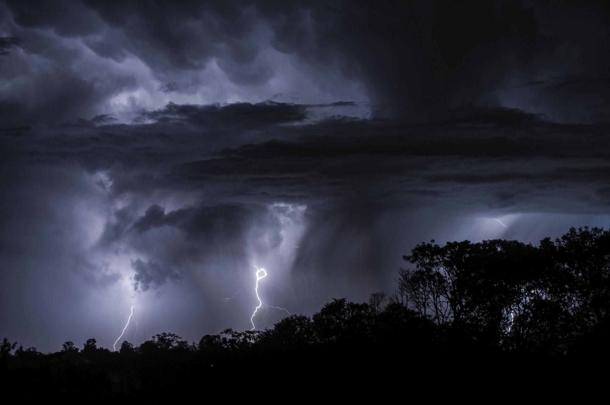 Lightning breaking through a massive dark thunderstorm above a silhouetted tree line