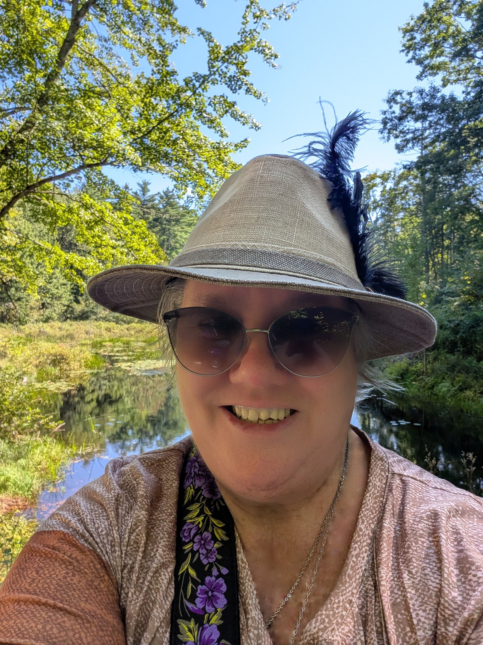 M. R. Brown smiling beside a woodland pond in a feathered hat